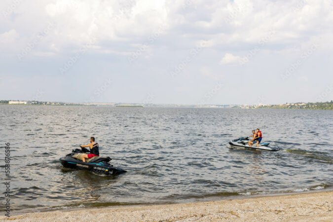 Nikolaev, Ukraine - June 18, 2021: Man and teenager coastguards use a jet-ski on the river coast.  Vacation Concept in the riverside at the city nature
